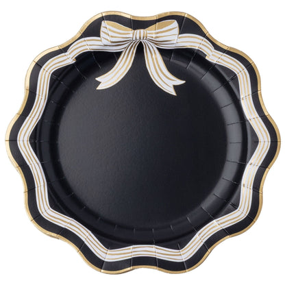 Round black paper party plates with a scalloped edge, featuring a decorative bow design in white and gold stripes along the top border.