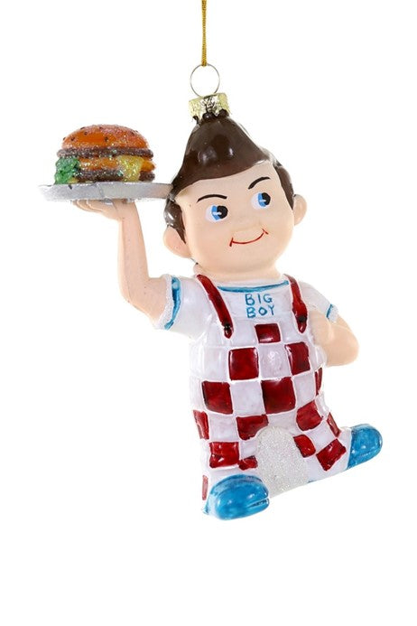 A glass ornament depicting a cartoon character resembling Bob's Big Boy, with a burger in one hand and a Santa hat in the other, dressed in a red and white checkered outfit.