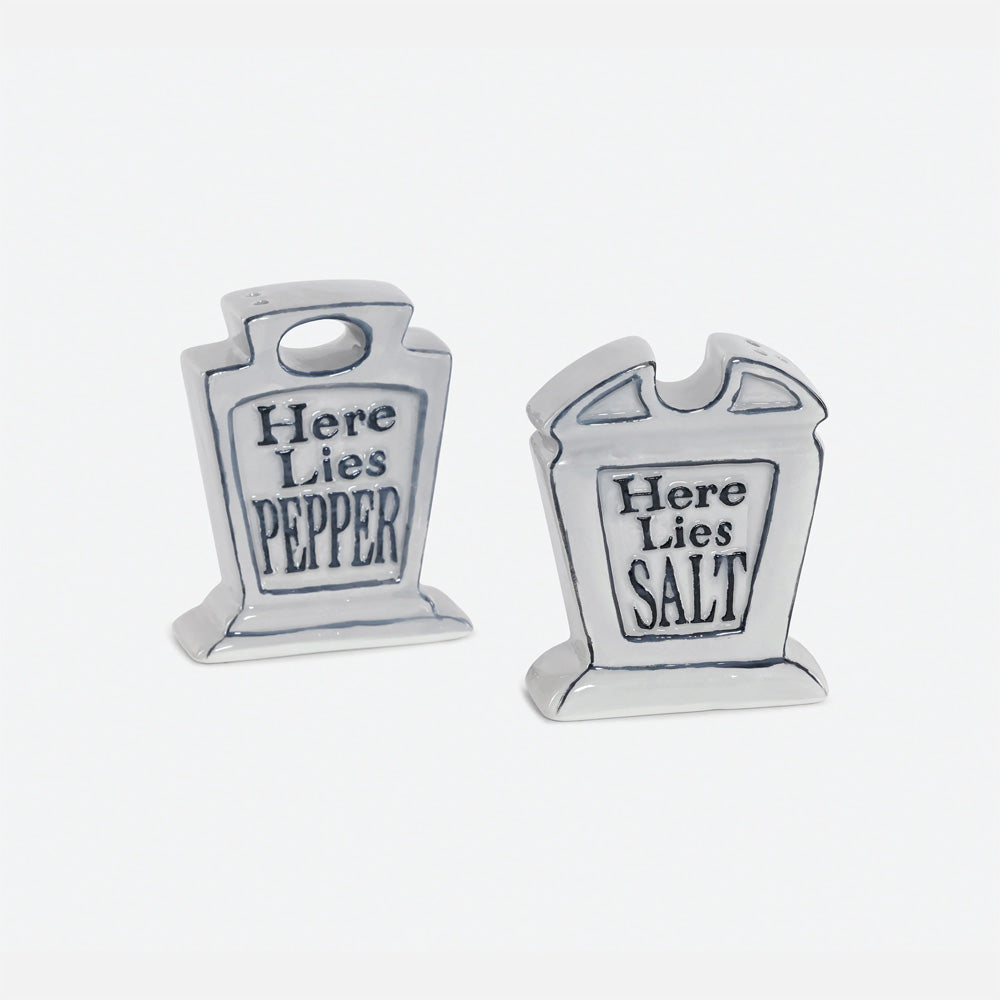 A pair of small tombstone-shaped salt and pepper shakers. One reads 'Here Lies PEPPER' and the other reads 'Here Lies SALT' in dark lettering.